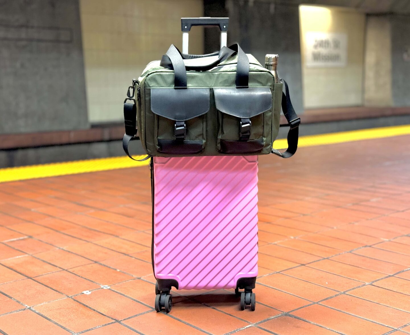 WaterField Designs X-Air Duffel in olive green sitting atop a bright pink rolling suitcase on a BART platform in San Francisco.